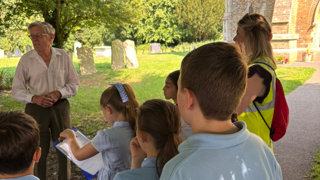 Primary school pupils carrying out fieldwork in High Ongar. The children are listening to a man speaking and writing things down.