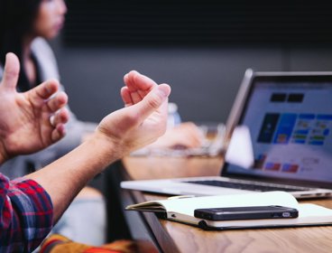 Person gesturing in a meeting with an open notebook, phone and laptop on a wooden table