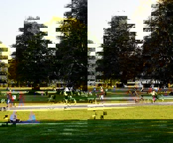 An area of parkland, showing people walking, sitting and cycling. It is a nice day and the sun is low in the sky which is casting long shadows from the trees.