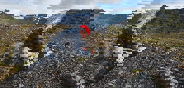 Researcher kneeling in a rocky landscape writing in a notebook. There is a glacier in the background.