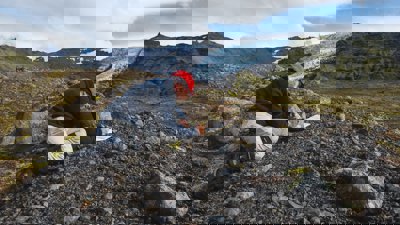 Researcher kneeling in a rocky landscape writing in a notebook. There is a glacier in the background.