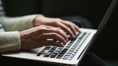 A persons hands typing on a computer. The background is dim and blurred.