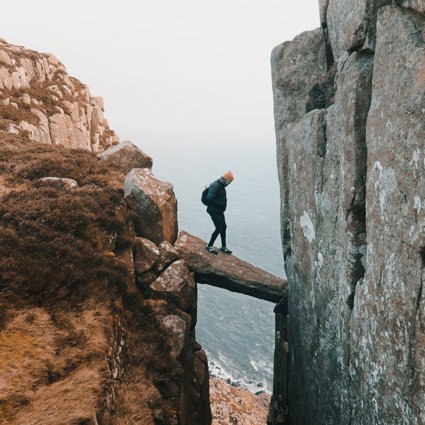 A person is standing on a precariously placed rock that is wedged between two cliff sides. They are high up and there is a long drop beneath the wedged rock onto a rocky beach.