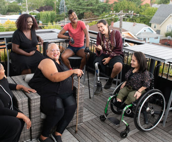 Six people sit down in chairs on a porch laughing. One person sits in a wheel chair, another holds a cain and another has a prosthetic leg.