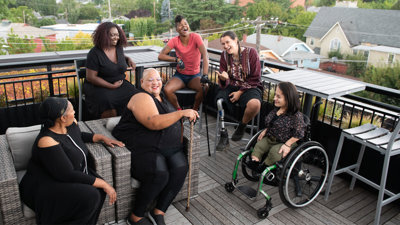 Six people sit down in chairs on a porch laughing. One person sits in a wheel chair, another holds a cain and another has a prosthetic leg.