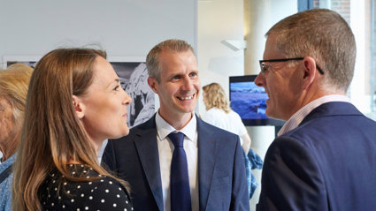 Three people in professional attire smiling and talking while standing in a bright exhibition space.