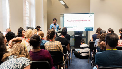 A presenter speaks to a room of people. A large screen show a slide of a PowerPoint presentation.
