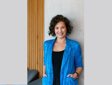 Person wearing a bright blue blazer standing with hands in pockets against a wall with wooden paneling.