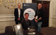 Three men in an office building smiling and posing for a photo in front of an Office for National Statistics Research Challenge Awards sign.