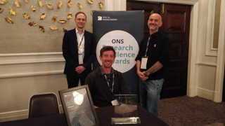 Three men in an office building smiling and posing for a photo in front of an Office for National Statistics Research Challenge Awards sign.