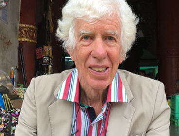 A man with white hair and wearing a colourful stripped shirt with a formal, beige jacket sitting outside a pagoda.