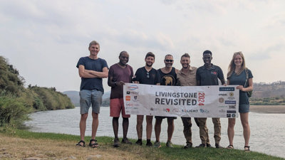 Group of seven people standing next to a river while holding a banner that reads 'Livingstone revisited 2024'.