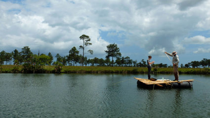 Two people stand on a wooden raft in the middle of a lake. The lake is surrounded by green grass land, sparsely dotted with trees.