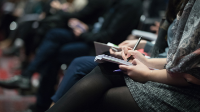Several people sit in the front row of what appears to be an auditorium or lecture hall. each person has a note pad and pen to hand, with some writing.